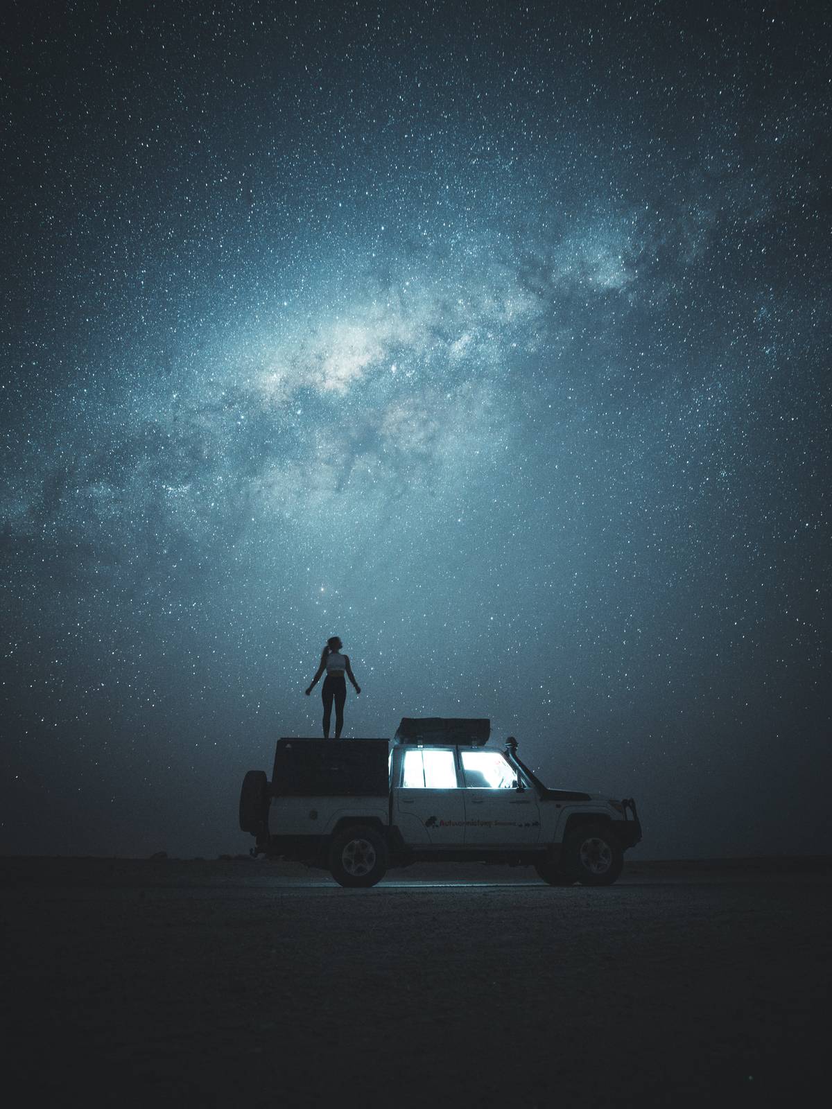 Solo photographer standing on a 4x4's rooftop tent under the Milky Way in a remote desert.