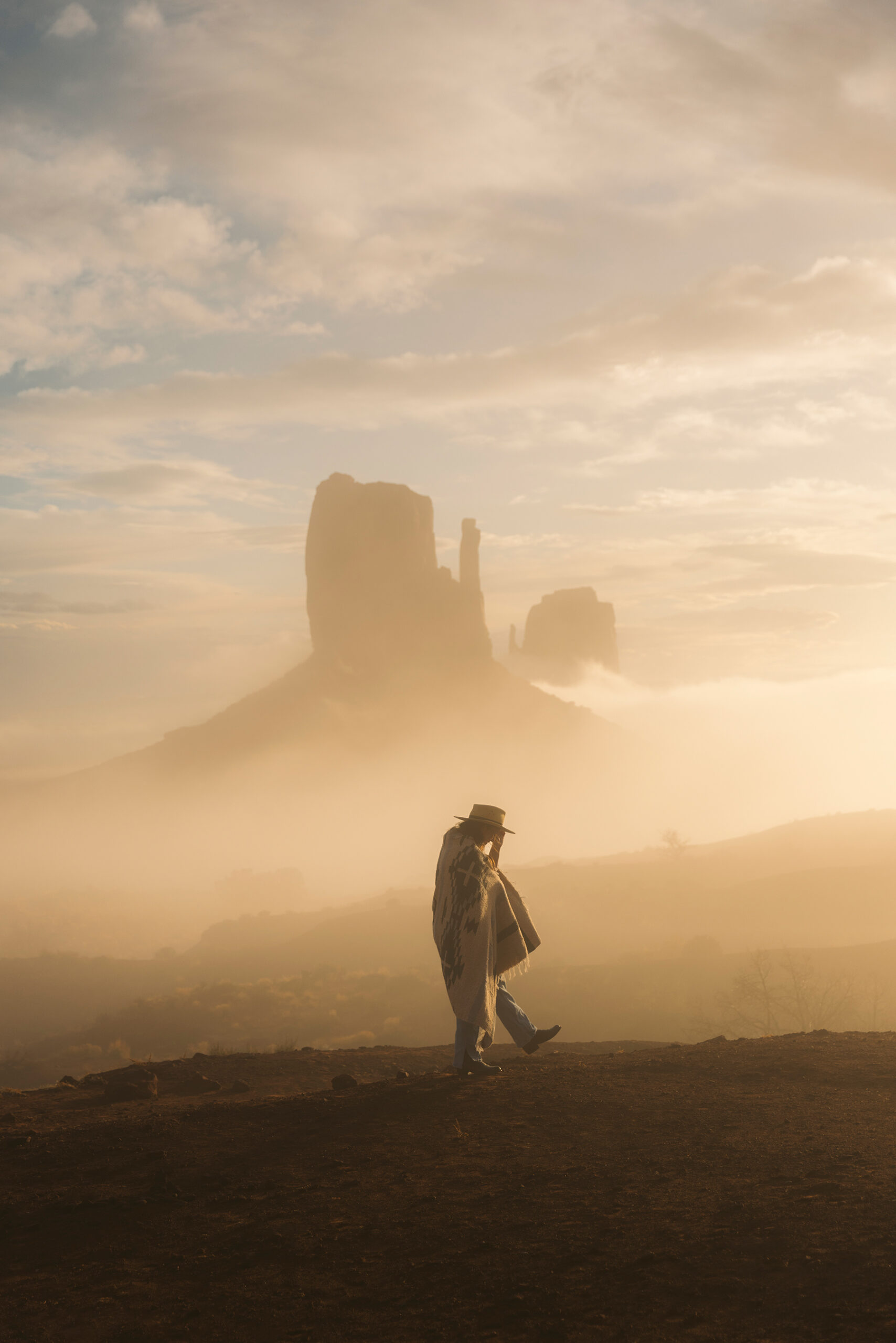 Conservation photography of a solo figure in a brimmed hat and patterned poncho walking across red earth as fog softens the sandstone buttes of Monument Valley in golden light.